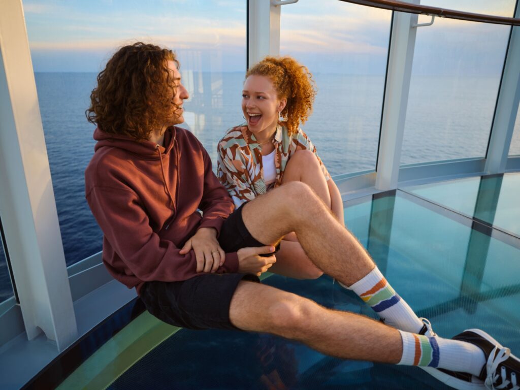 Zwei junge Erwachsene sitzen auf dem Skywalk von AIDA Cruises mit dem Meer im Hintergrund.